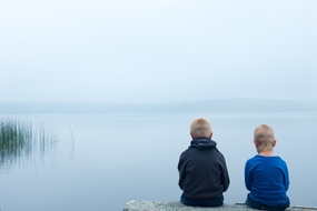 Illustrasjonsfoto: To barn sitter ved sjøen, med ryggen vendt mot kamera